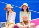 Two young women sitting on a tennis court wearing hats.